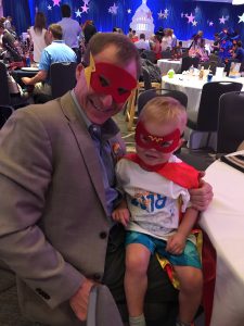 A young boy in a red superhero mask is on the lap of a man in a suit and a superhero mask