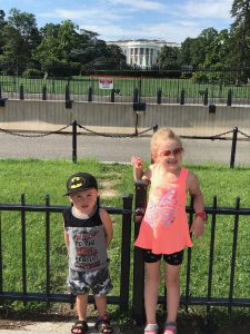 A small boy in a batman hat and a young girl in a pink tank top standing in front of the white house